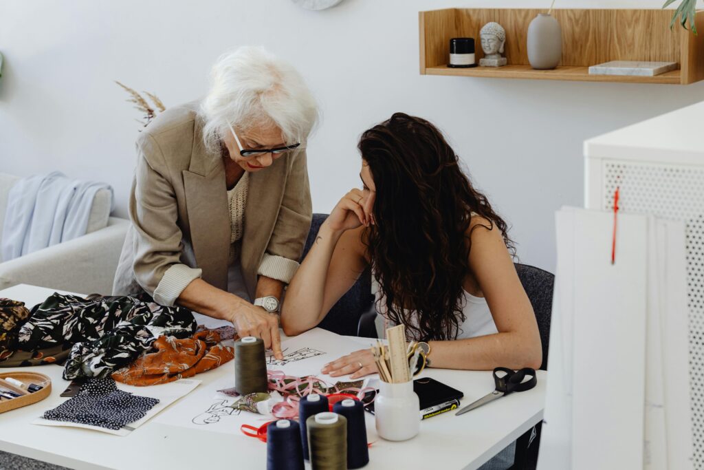 Elderly woman instructs young designer at worktable, sharing expertise in a fashion studio.